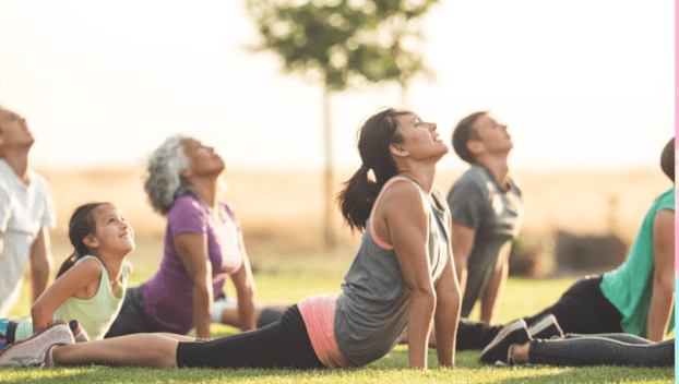 People of all ages do a yoga pose outdoors in a park.